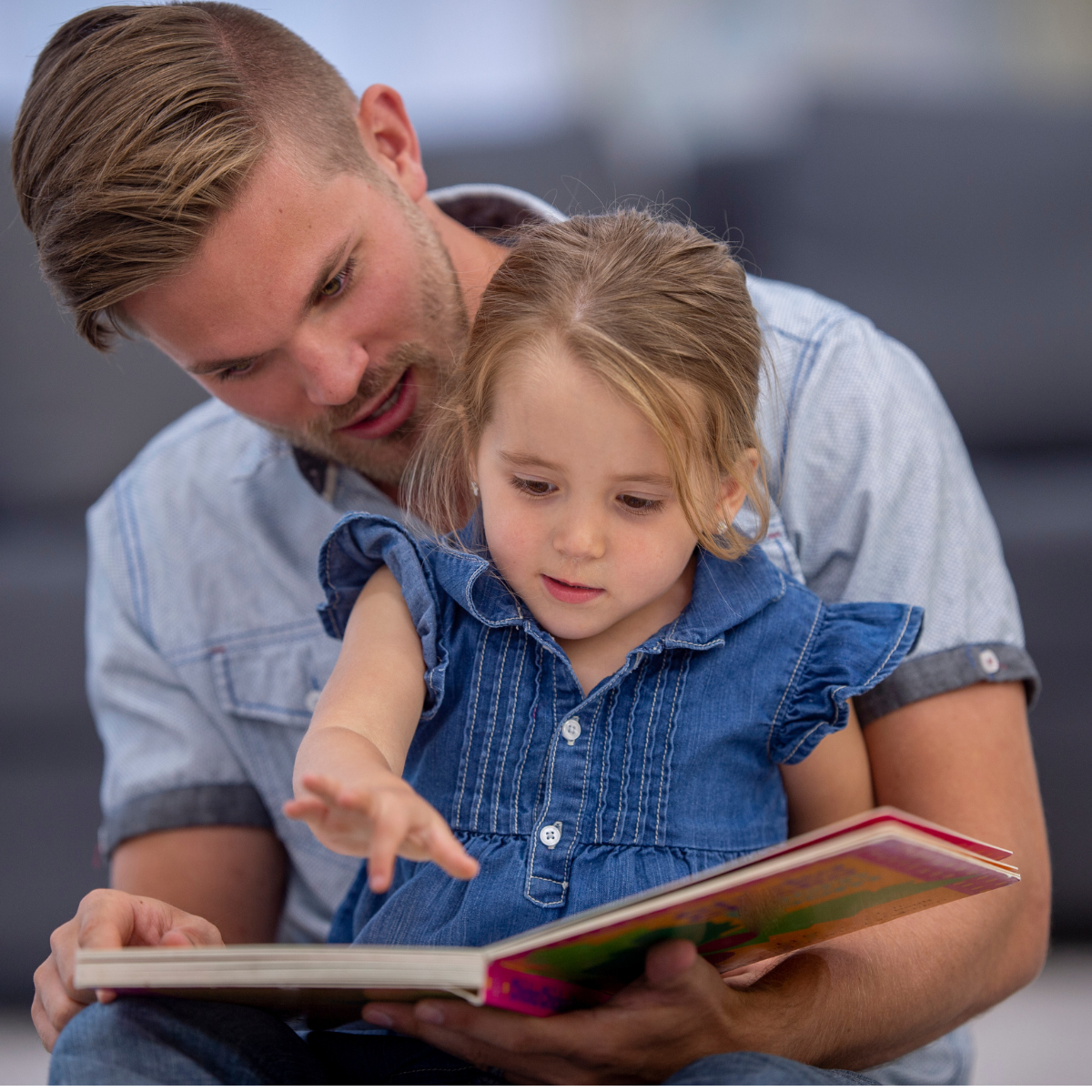 Ein Vater liest seinem kleinen Kind ein Buch vor. Das Kind sitzt auf seinem Schoß, beide schauen aufmerksam auf die Buchseiten.