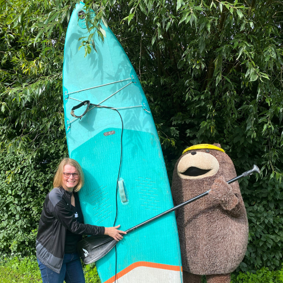 Anja Prem mit dem Grömel-Maskottchen neben einem SUP-Board.